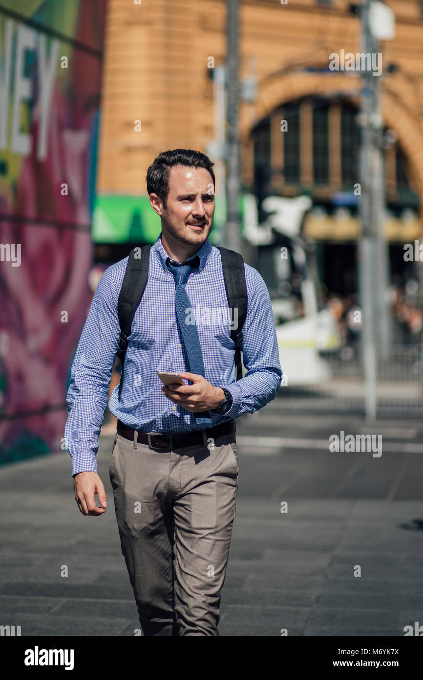 Millennial businessman is commuting to work in Melbourne, Victoria. He is walking while holding a smartphone. Stock Photo