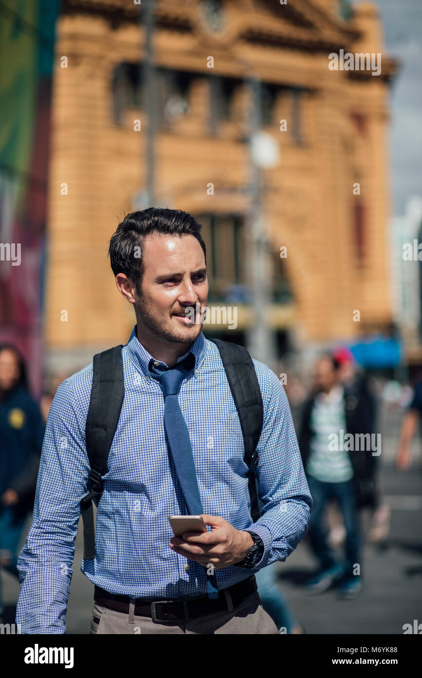 Millennial businessman is commuting to work in Melbourne, Victoria. He is walking while holding a smartphone. Stock Photo