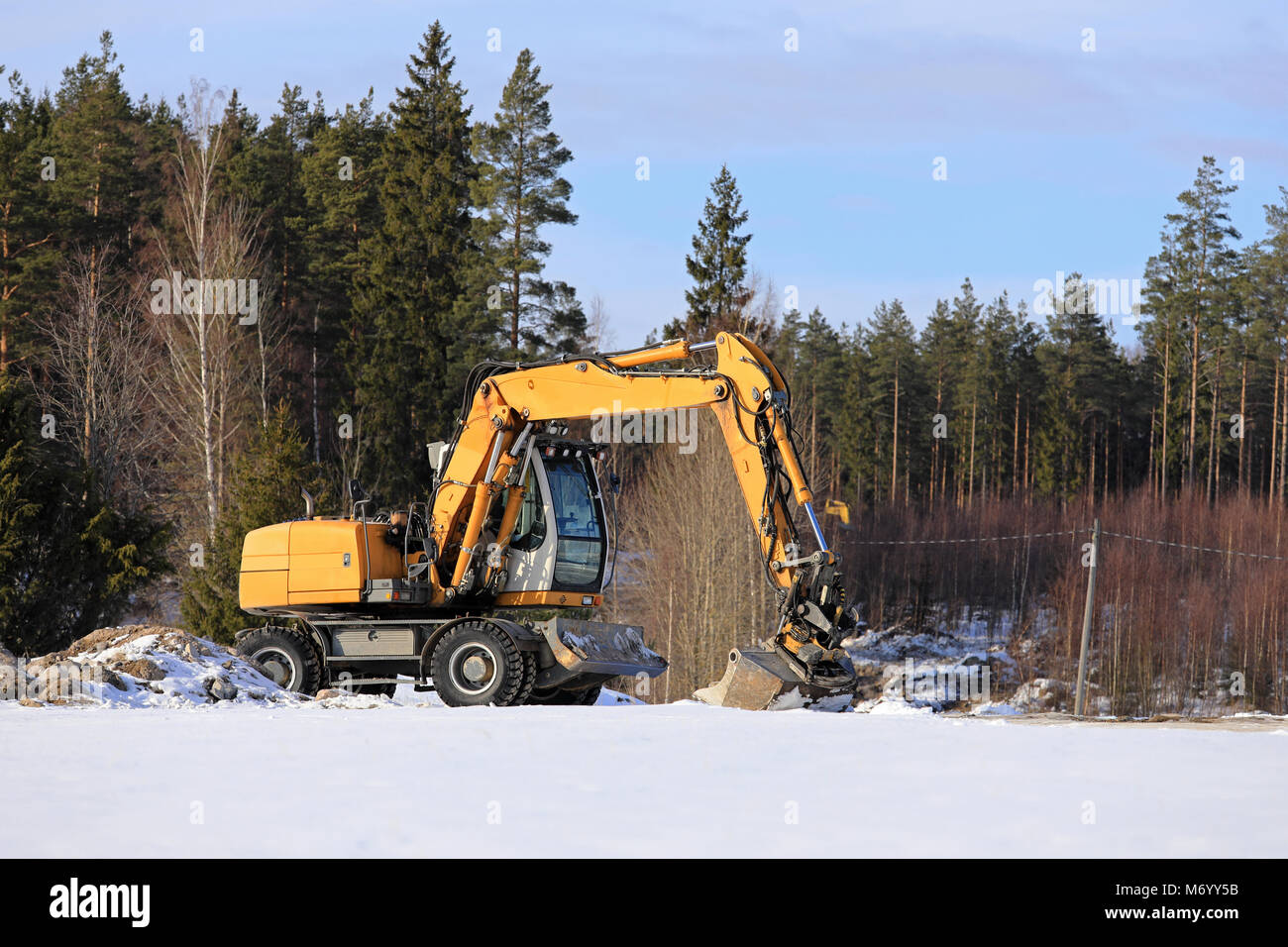 Yellow mid-size hydraulic excavator at rural construction site with forest on the background on a sunny day of winter. Stock Photo