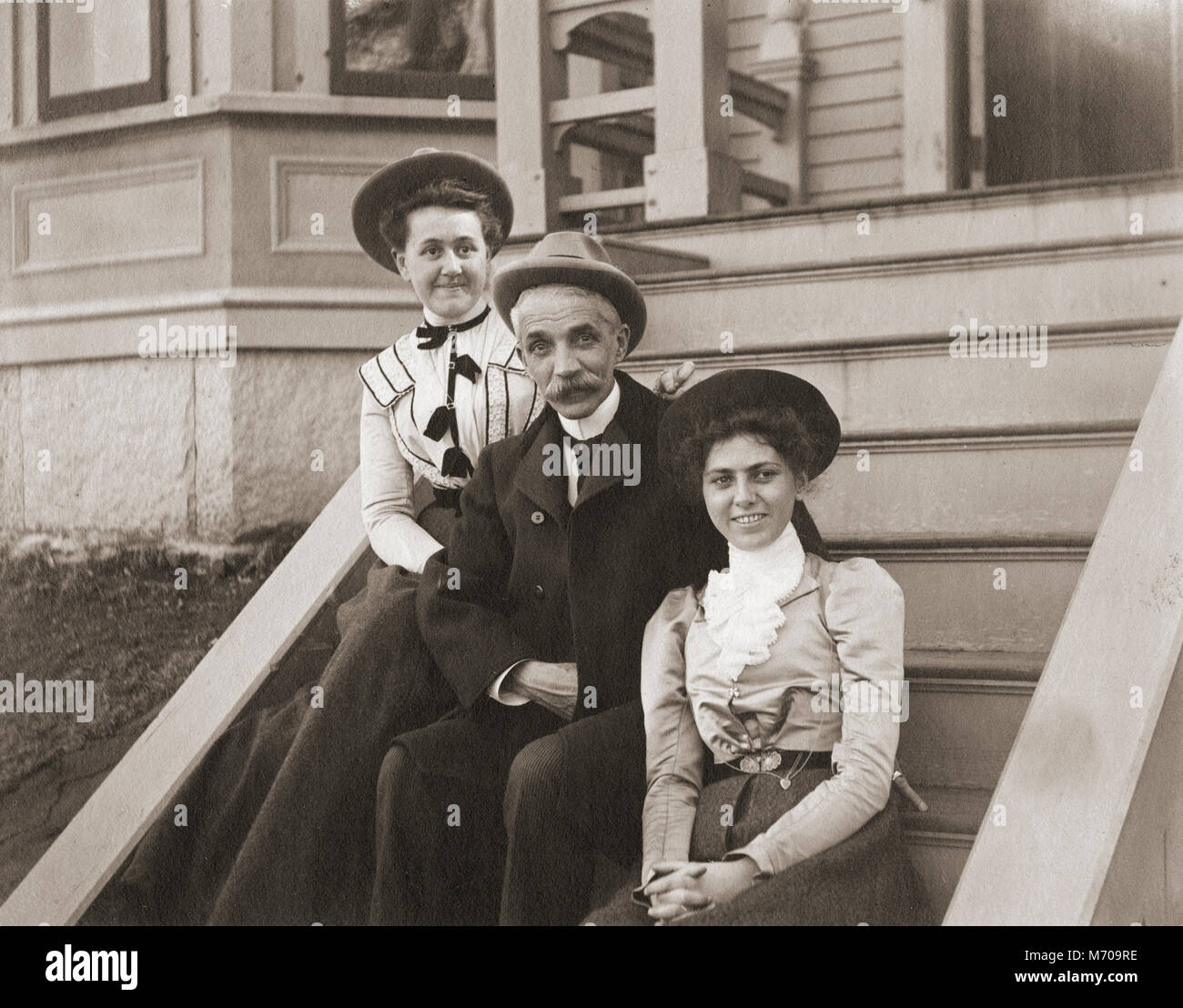 Antique circa 1905 photograph, father and two grown daughters sitting on front stairs of their home. Location unknown, probably New England, USA. I have more photos of this family in Riggsville, Maine, USA. Stock Photo