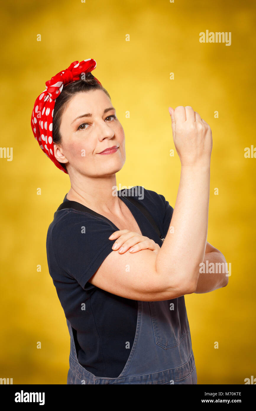 Self-confident middle aged woman with dungarees rolling up her sleeve, text space, tribute to american worker icon Rosie Riveter Stock Photo