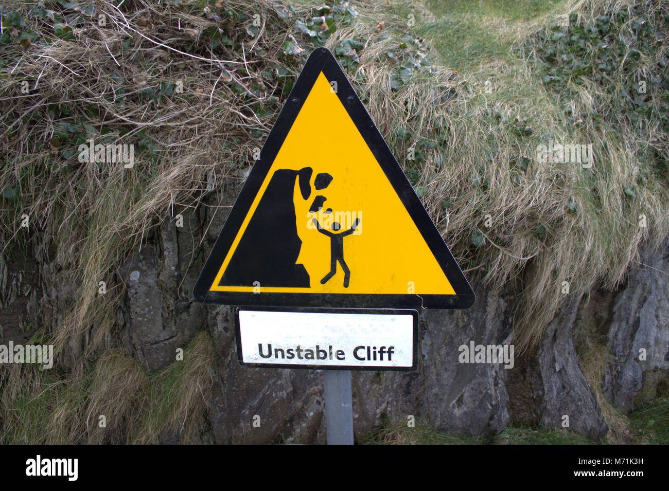 Danger from falling rocks sign at cliff face, which overhangs a narrow path down to a popular bathing spot. Stock Photo