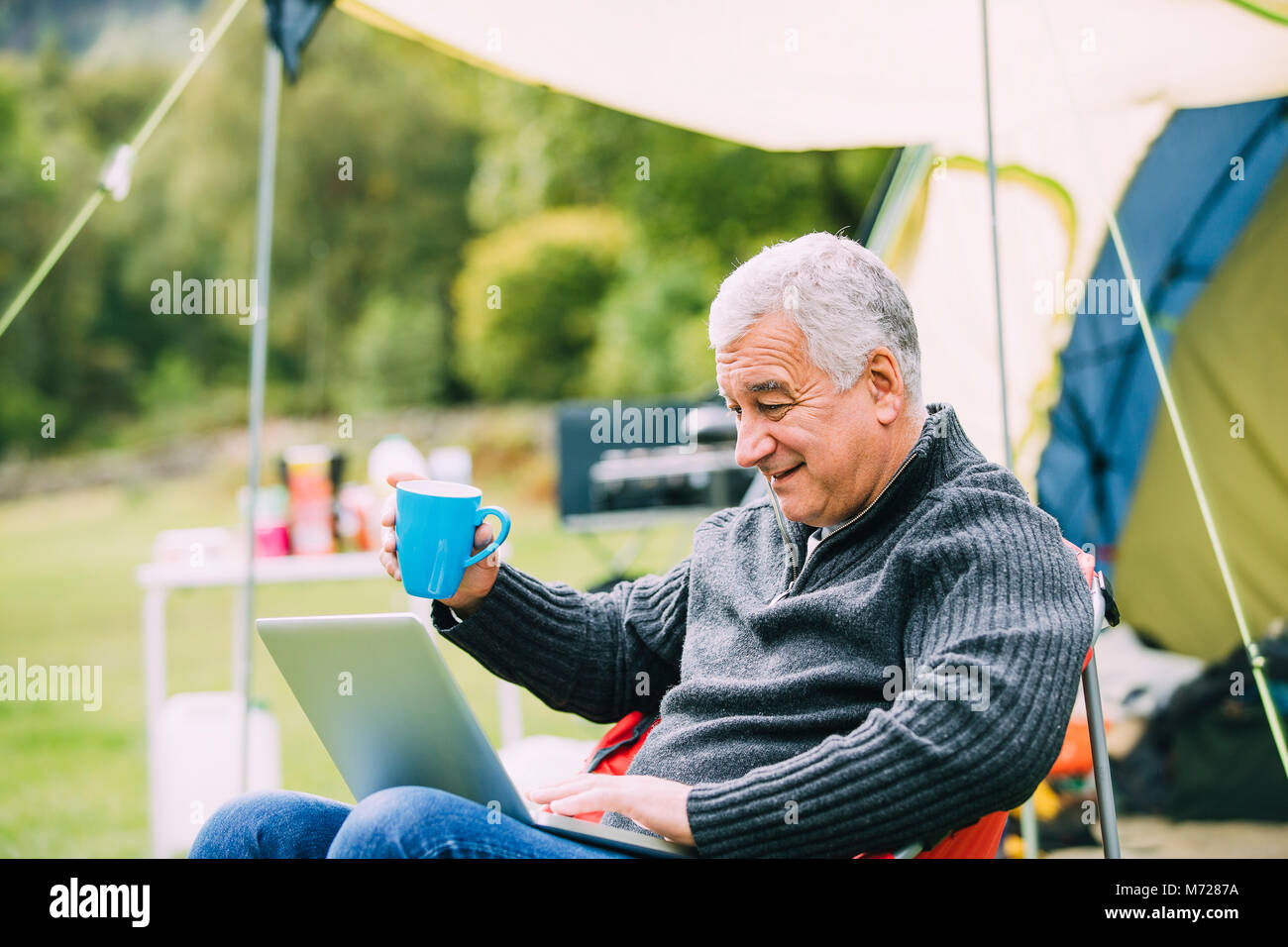 Senior man is relaxing in a chair by his tent in the campsite. He is enjoying a cup of tea while using a laptop. Stock Photo