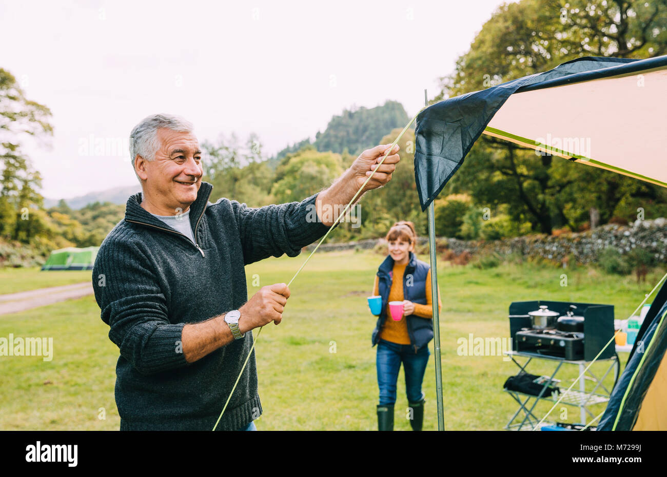 Senior couple have arrive at the campsite they are staing in. The man is putting up their tent while his wife makes cups of tea for everybody. Stock Photo