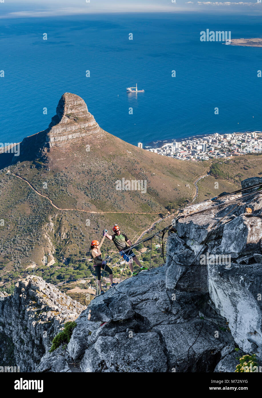 A couple about to abseil from the summit of Table Mountain in Cape Town. Lion’s Head, Robben island and Cape Town’s Atlantic seaboard can be seen in t Stock Photo