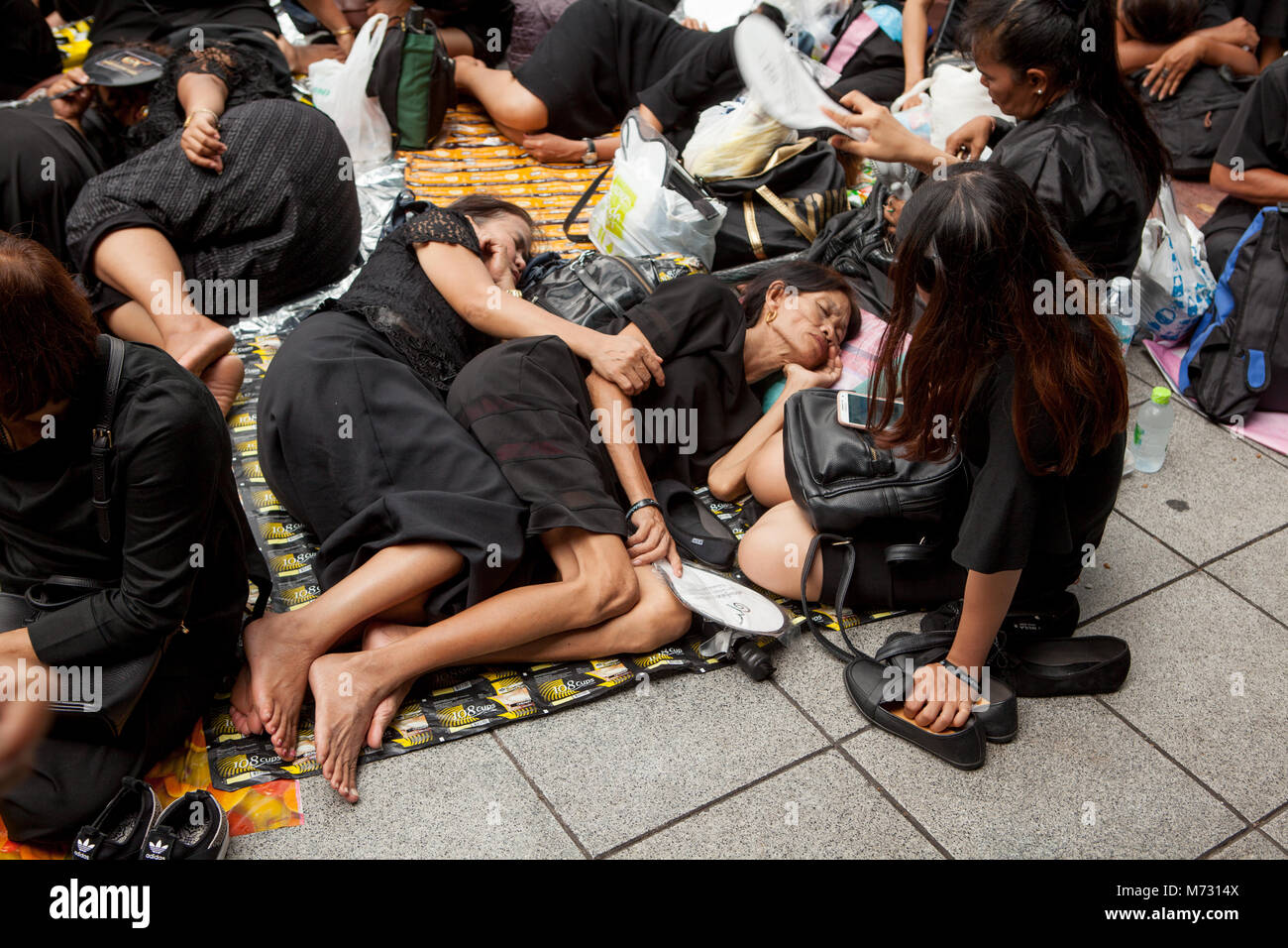 Two women sleeping in the street and hugging each other, in a crowd of people in black clothes, waiting for the Royal Cremation in Sanam Luang Stock Photo