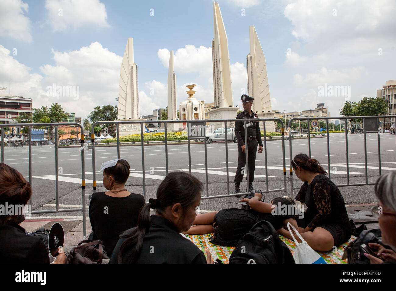 Thai police officer and women in black clothes sitting on the sidewalk in front of the Democracy Monument in Bangkok during the Royal Cremation Stock Photo