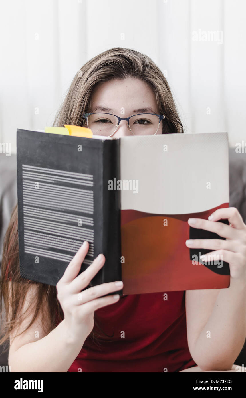 Woman holding a book in front of her with just the eyes out of the book. Student woman wearing glasses, Brazilian, japanese descendant. Stock Photo