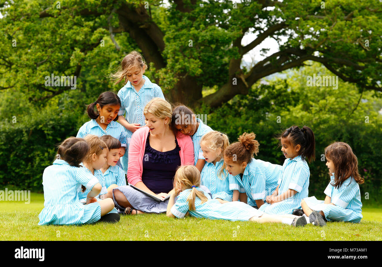 Primary schoolchildren gathered around their teacher outside under an oak tree reading a story Stock Photo