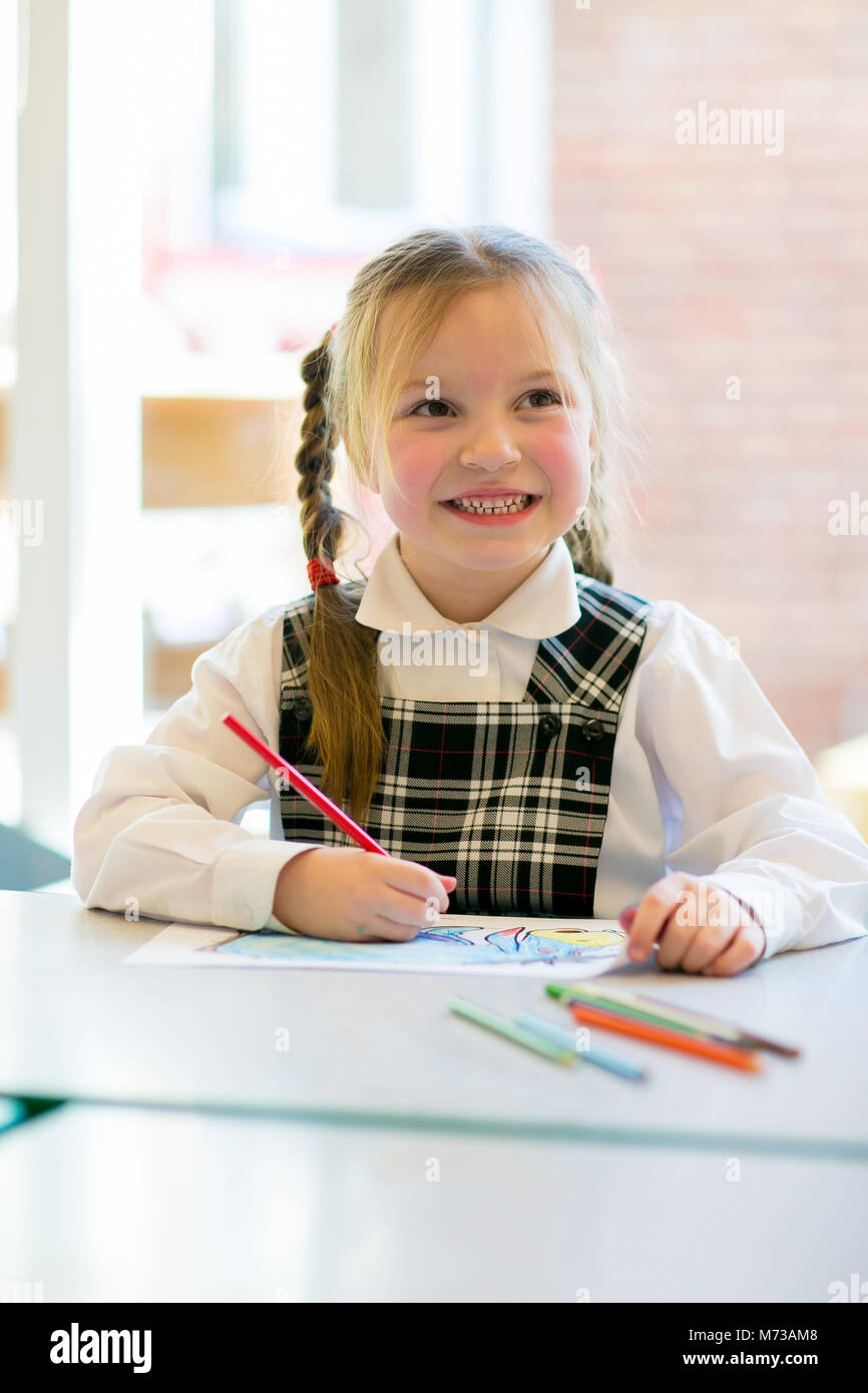 Primary schoolgirl sitting at a low table drawing, looking up smiling with a pencil in her hand Stock Photo