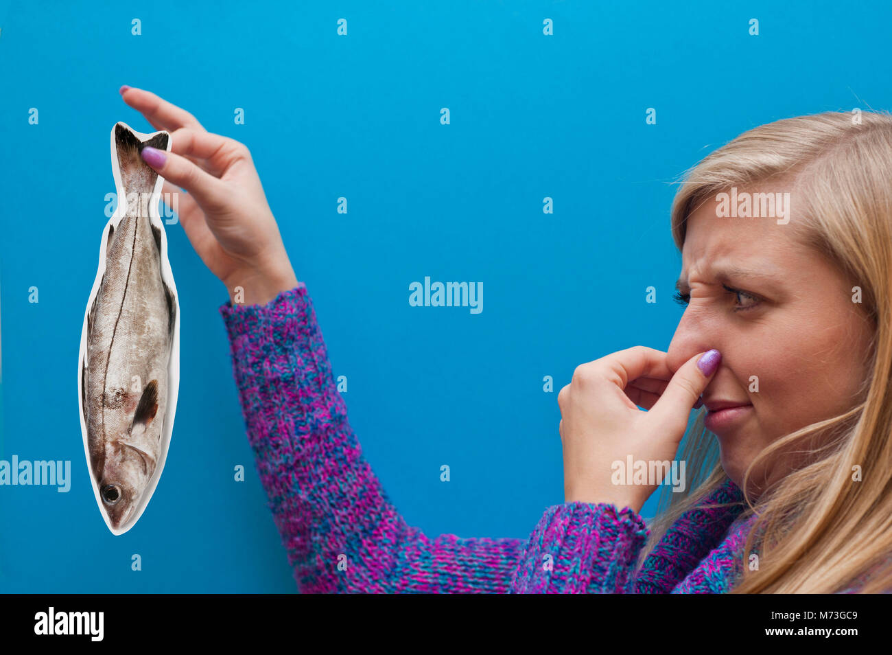 blonde young woman keeping a fake card fish and keeping her nose in disgust, concept of English saying: 'smells fishy' - somebody being dishonest. Stock Photo