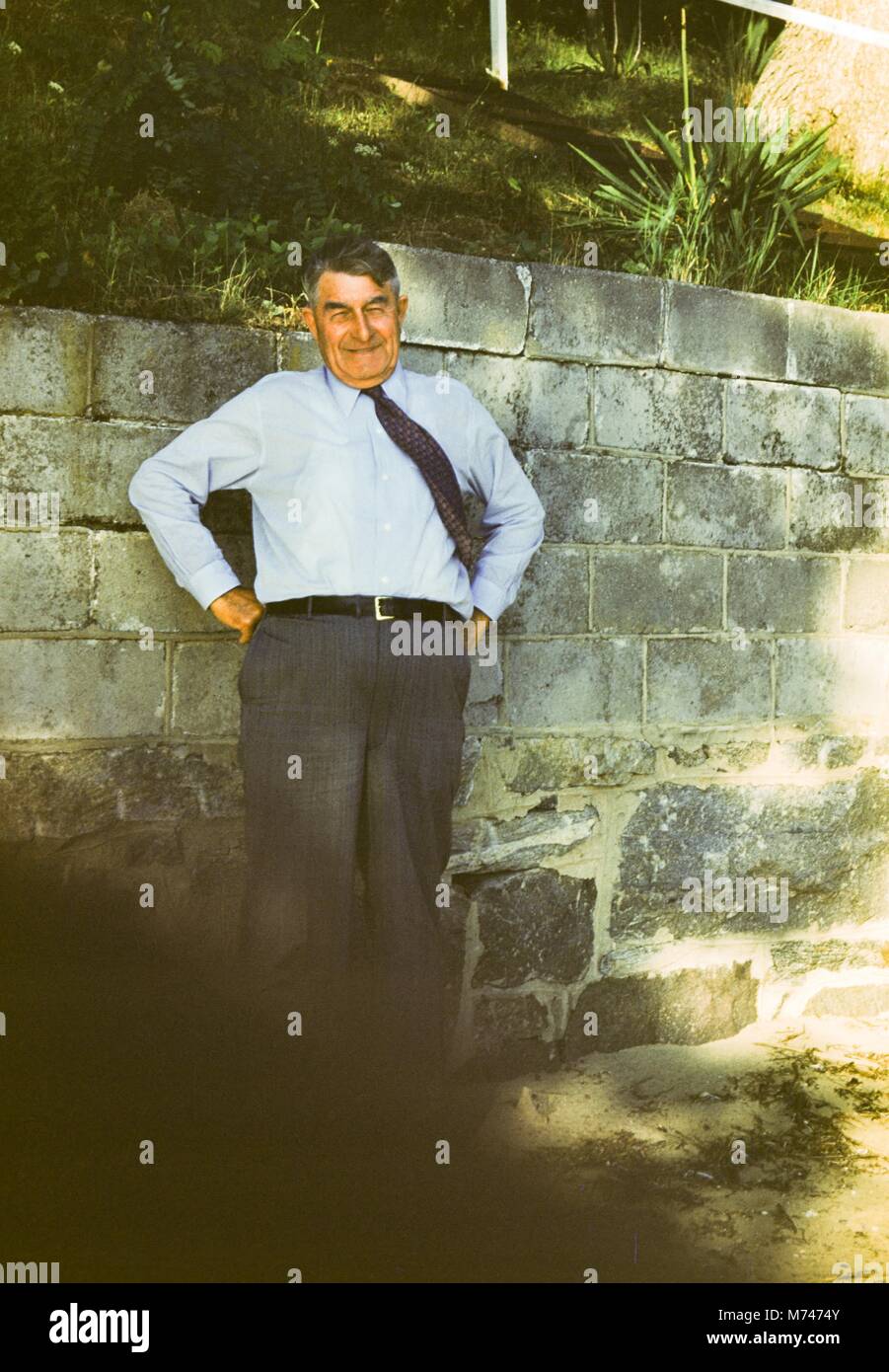 A mature man wearing dress clothes and a tie poses with his hands on his hips by a concrete retaining wall, 1955. () Stock Photo