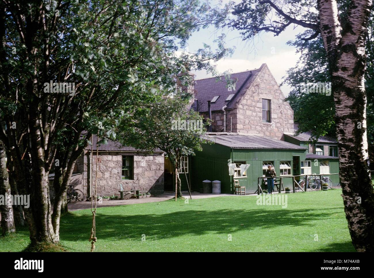 A woman stands near a roughly constructed wooden fence outside of a large stonework home with wooden additions, 1965. () Stock Photo