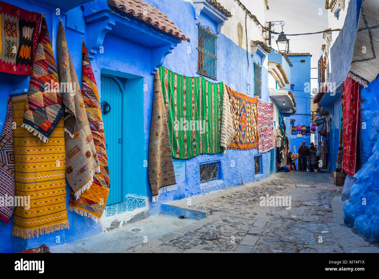 A colourful narrow street scene in the blue city of Chefchaouen, Morocco. Rugs, mats carpets on display in cobbled street with people in distant view Stock Photo