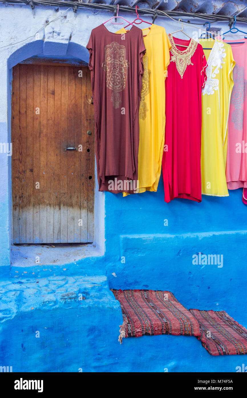 red coloured rug on steps leading to a door and colourful traditional dresses clothing hanging in a street in Chefchaouen, Morocco Stock Photo
