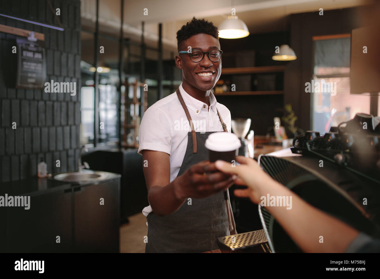 Coffee shop owner handing over a sealed coffee cup to a customer. Man serving customer with a smile at a coffee shop. Stock Photo