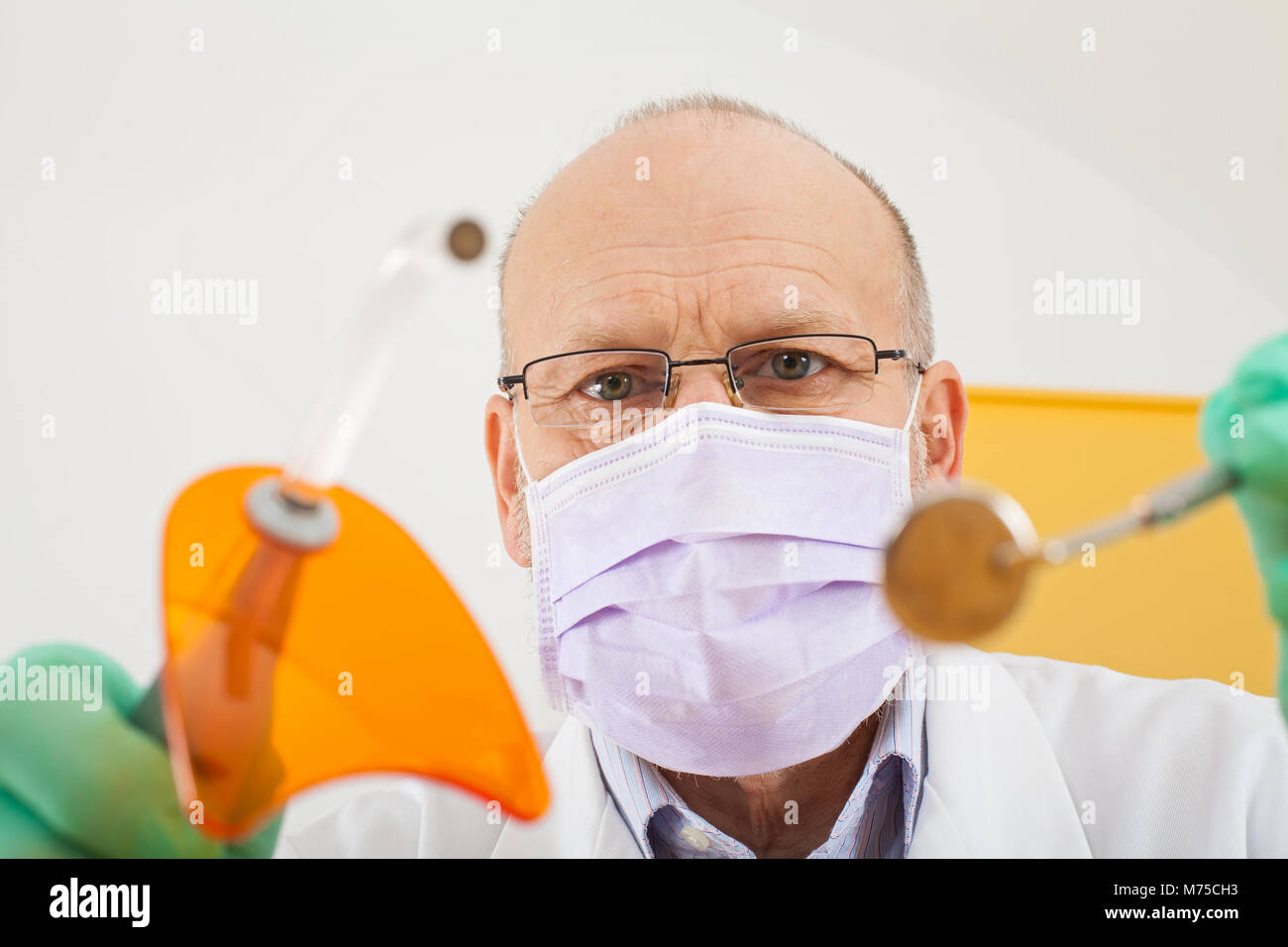 Mature male dentist wearing mask holding dental instruments, looking down at the camera Stock