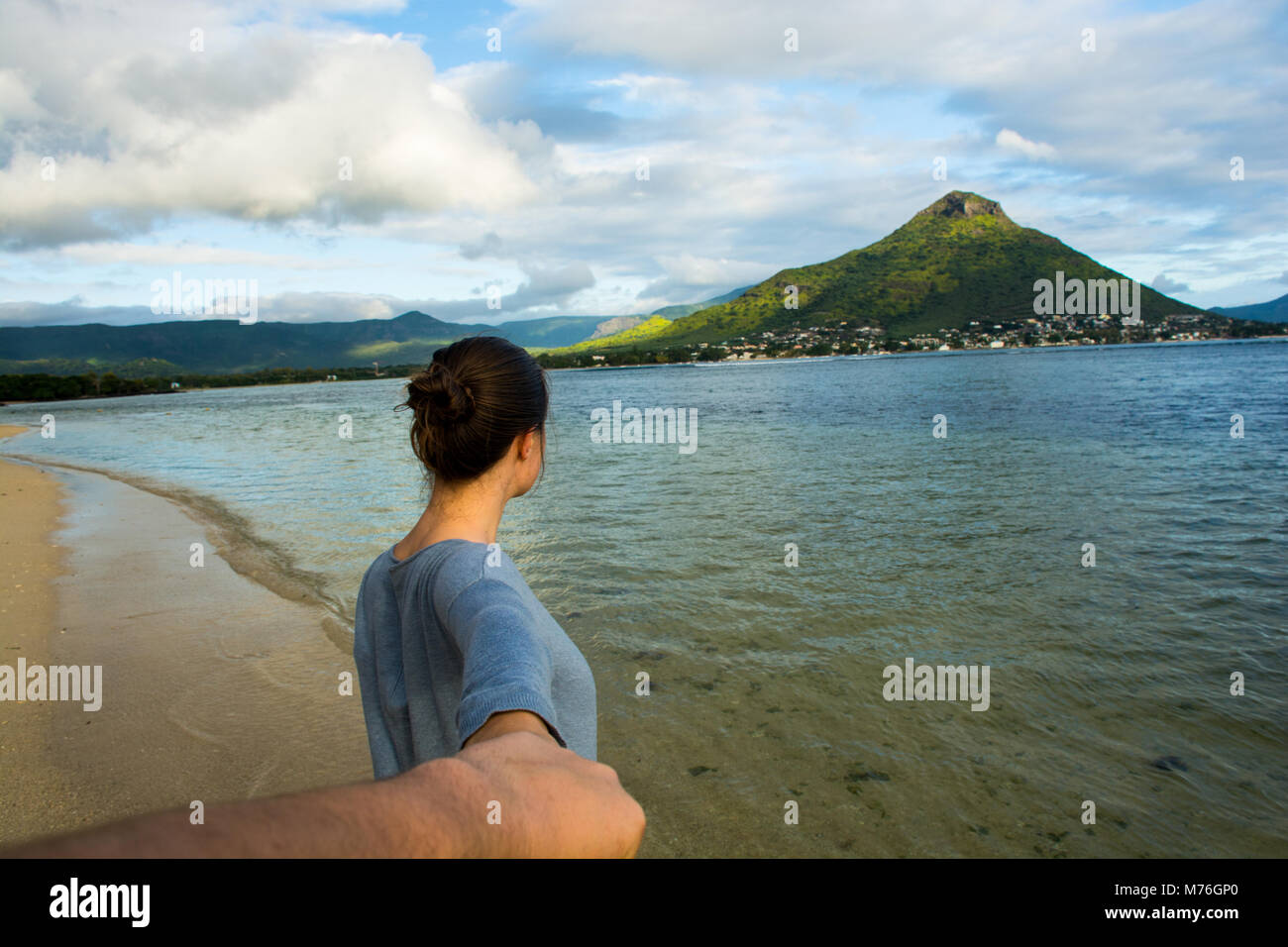 This young woman is leading the way for her partner, walking towards the ocean with the mountains in the background. Stock Photo