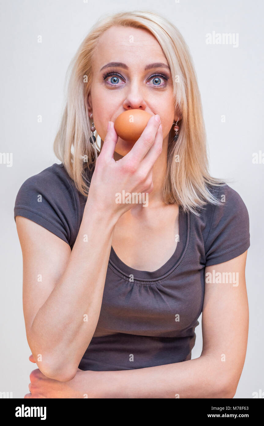 Big eyes young blonde woman holding chicken egg near her mouth on white background Stock Photo