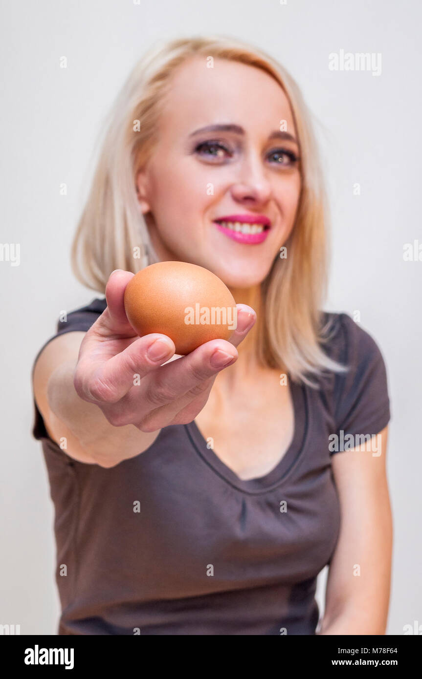 Young smiling blonde woman holding showing offering brown chicken egg on white background Stock Photo
