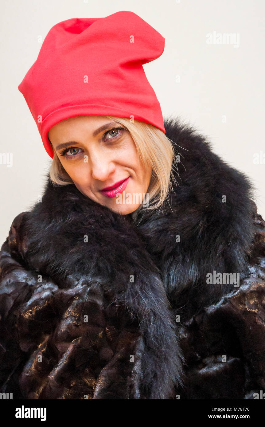 Portrait of young blonde smiling woman with red hat in black fur coat. Stock Photo