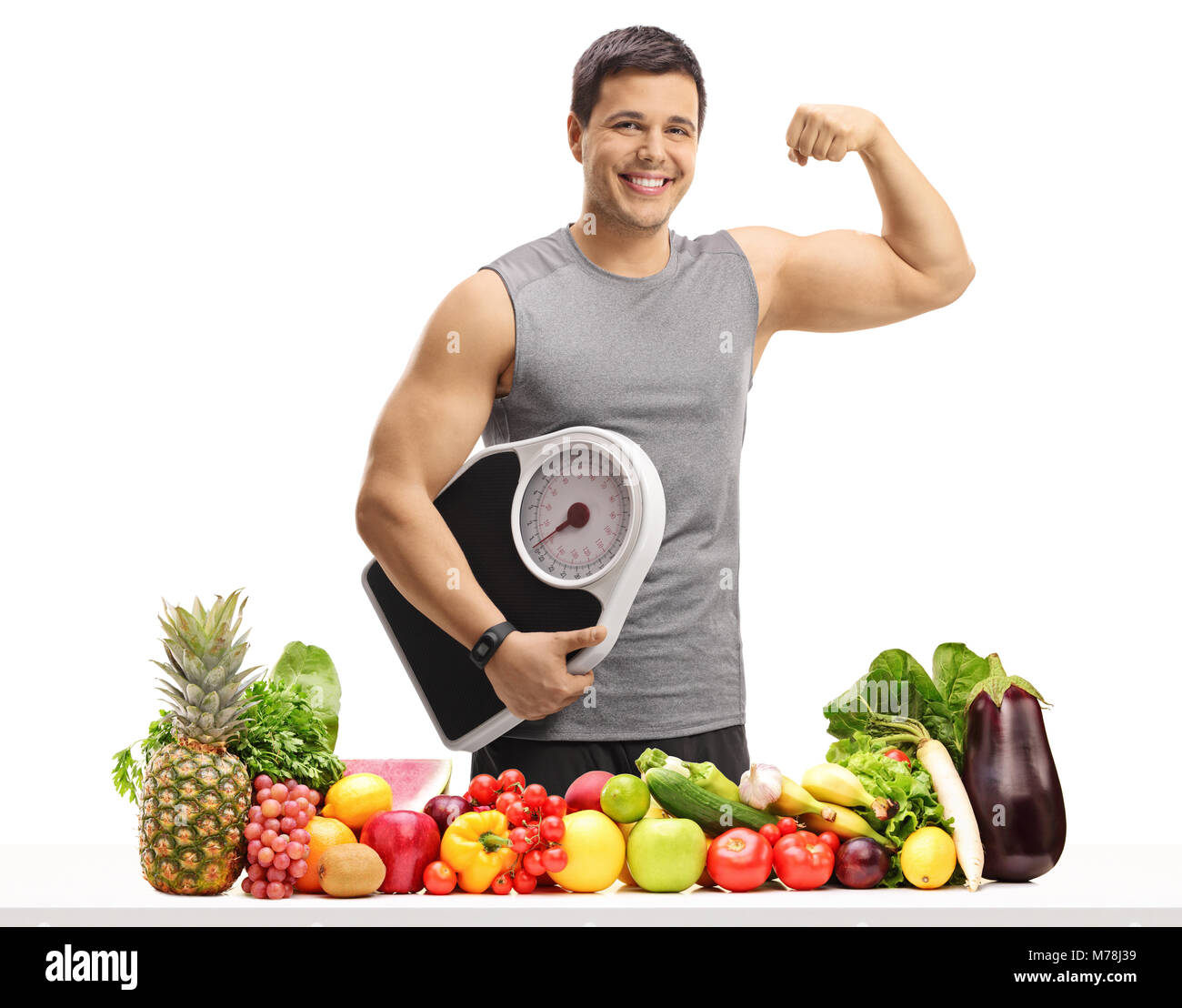 Young man in sportswear holding a weight scale and flexing his biceps behind a table with fruit and vegetables isolated on white background Stock Photo