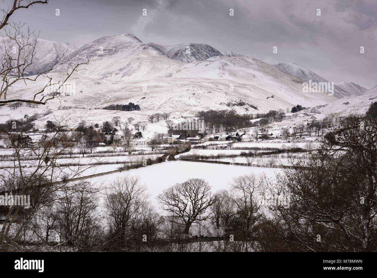 Bleak grey sky over a cold winter view of snow covered Buttermere village with the snowy fells Whiteless Pike and Wandope beyond Stock Photo