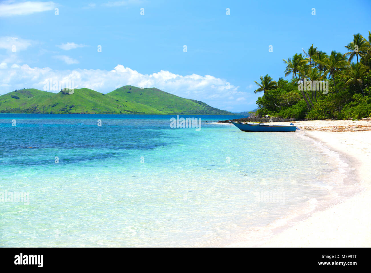 A small dark blue boat parked at white sandy beach on tropical Fiji island. Green hills in background and coconut palm trees behind the boat. Stock Photo