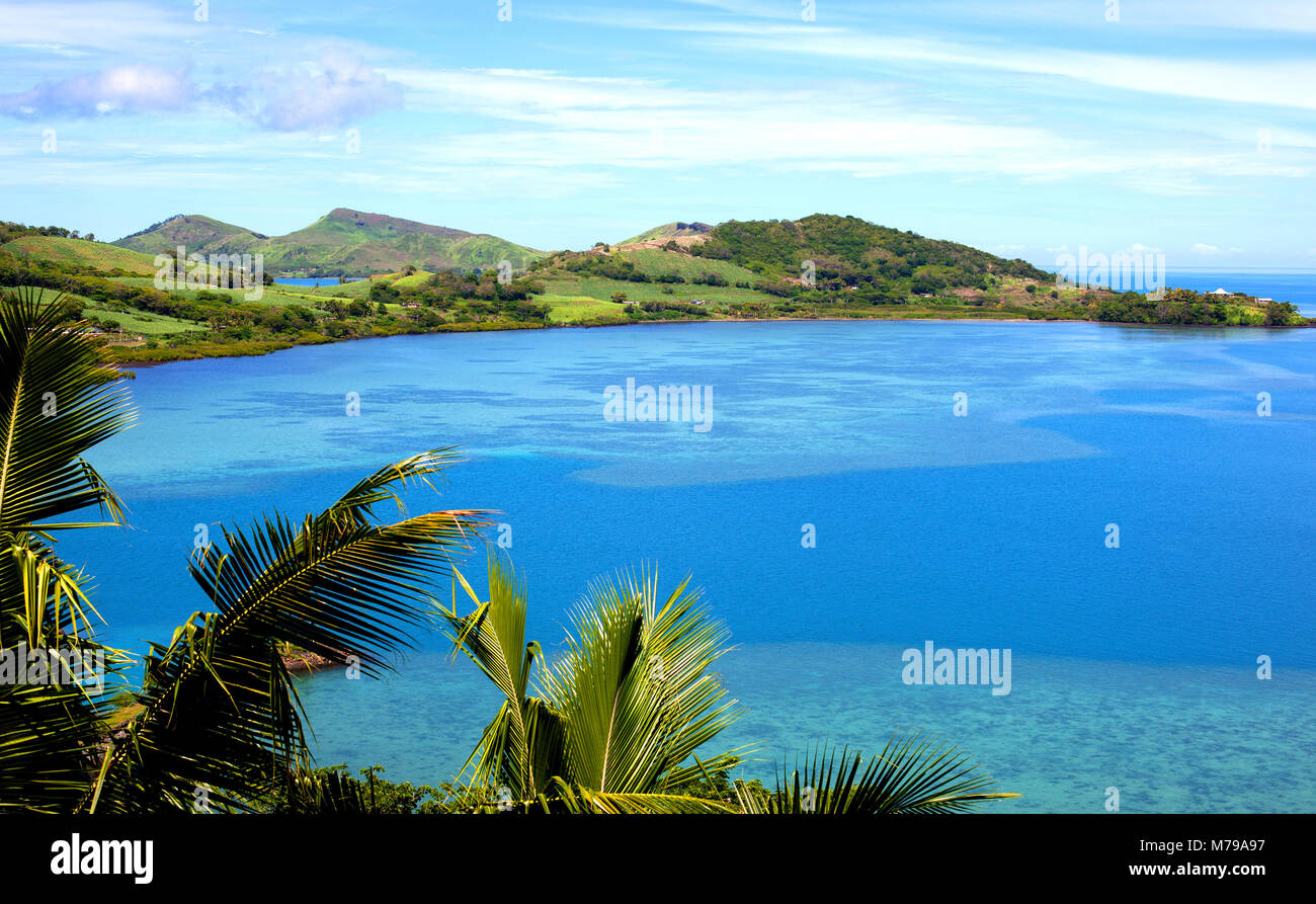 Beautiful landscape of Fiji island during high tide with palm trees in foreground and ocean with green hills in background during sunny day. Stock Photo