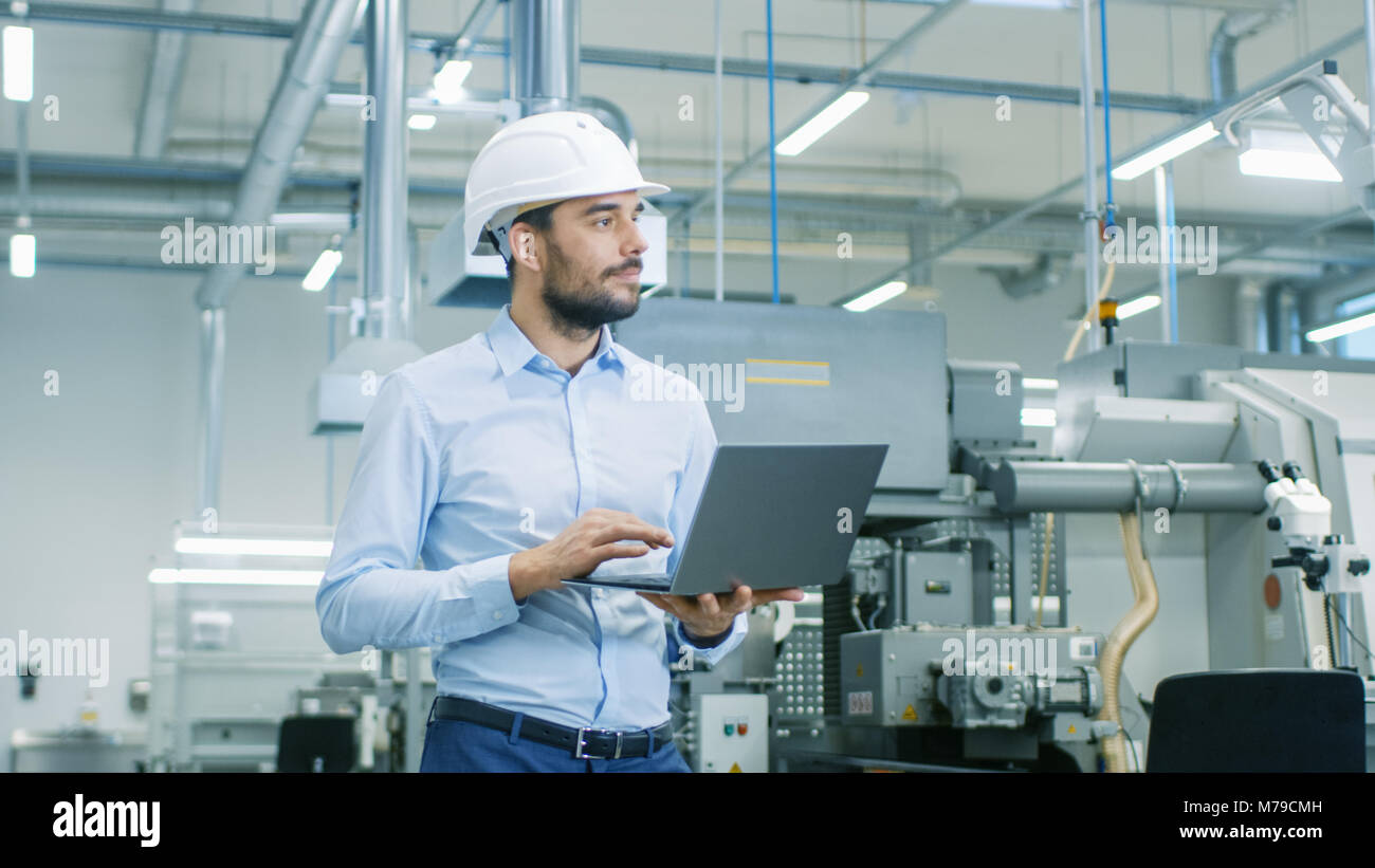 Chief Engineer in the Hard Hat Walks Through Light Modern Factory While Holding Laptop. Successful, Handsome Man in Modern Industrial Environment. Stock Photo