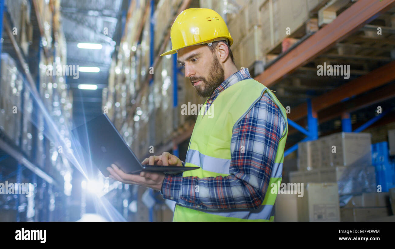 Warehouse Worker Uses Laptop. He's Standing in the Middle of  a Big Distribution Center with Big Storage Racks and Pallets on Them. Stock Photo