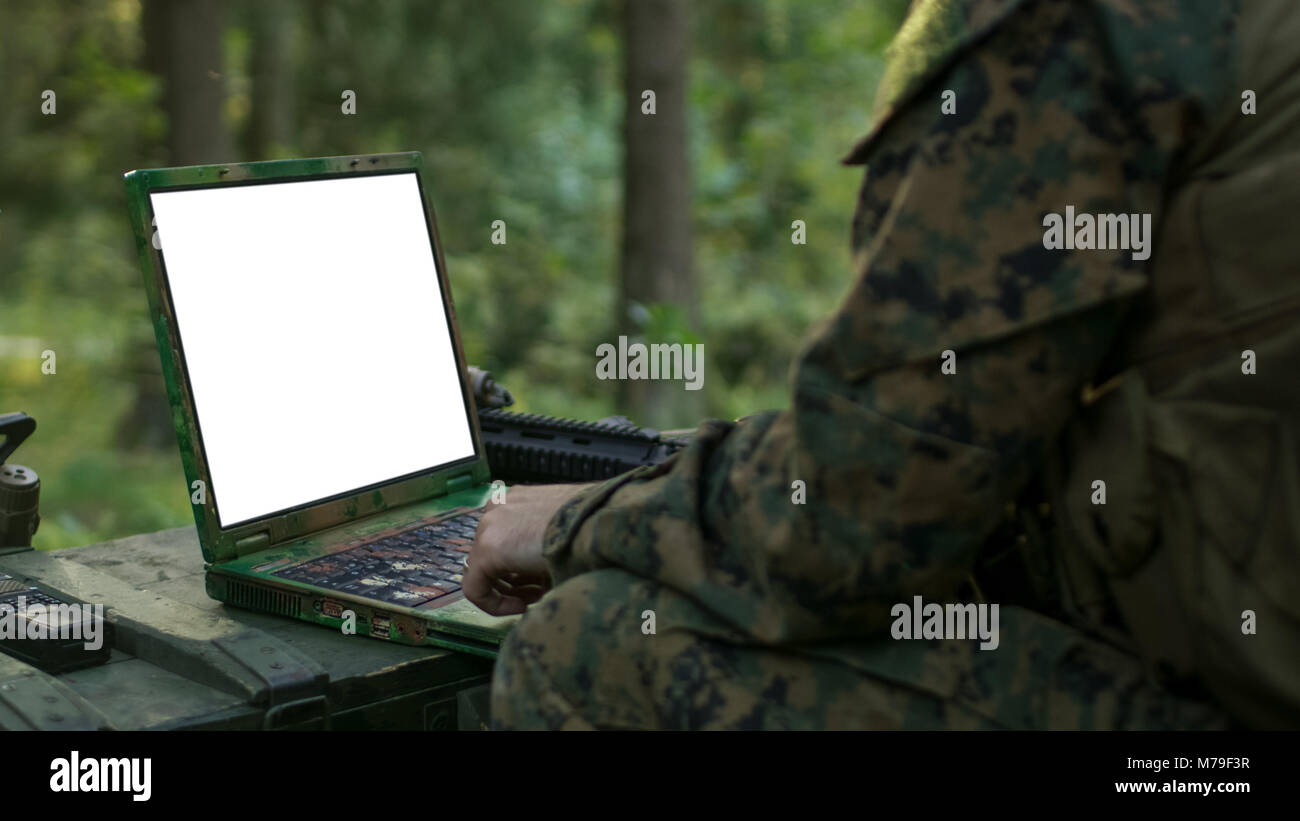 Close-up Soldier Uses Military Grade Laptop with White/Green Screen. In the Background Camouflaged Army Base in the Forest. Stock Photo