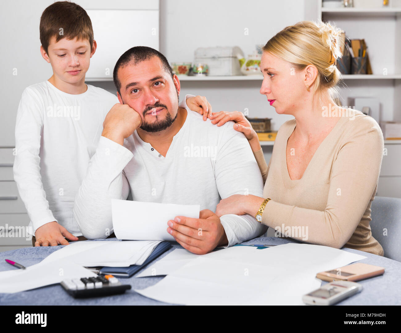 Chagrined man sitting at home table with papers while family soothing ...