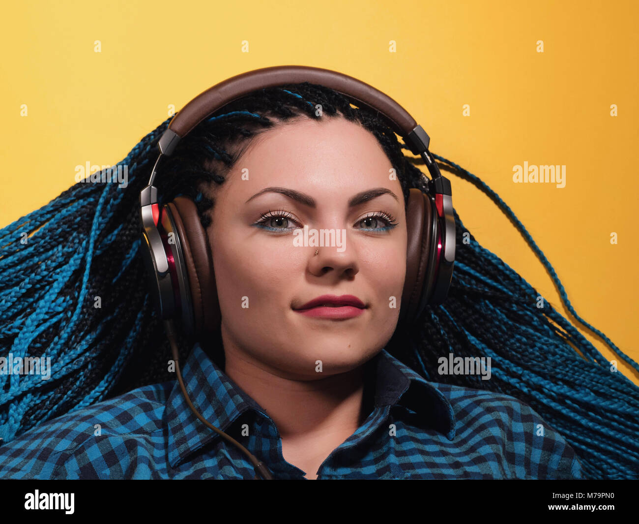 Beautiful young girl with african blue braids is listening to music with headphones and dancing. Woman on bright yellow background. Dyed Hair moves. Stock Photo