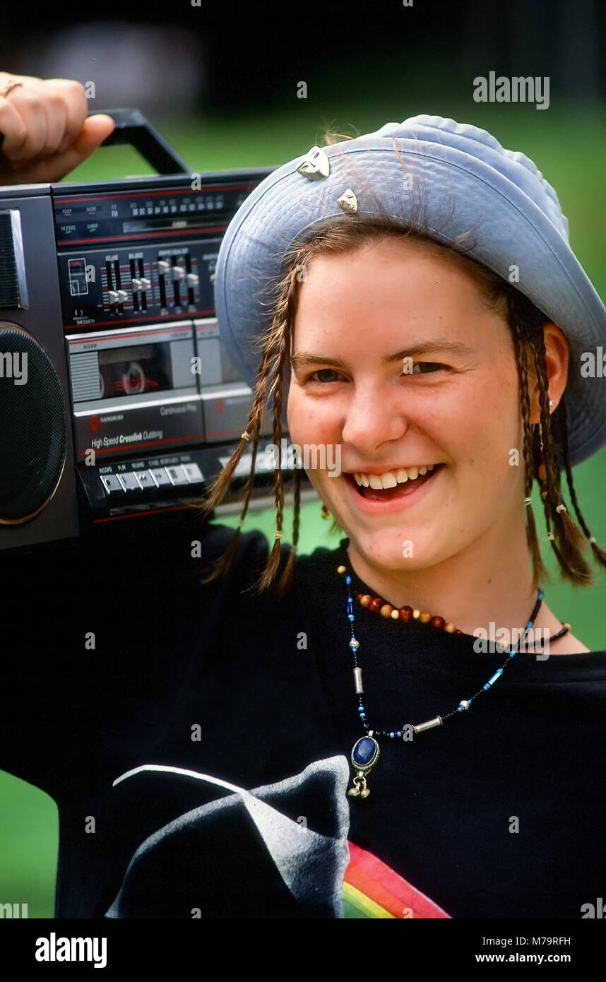 A teenage girl with braided hair, blue hat, necklace and black shirt carries her boombox on her shoulder at summer camp. Stock Photo