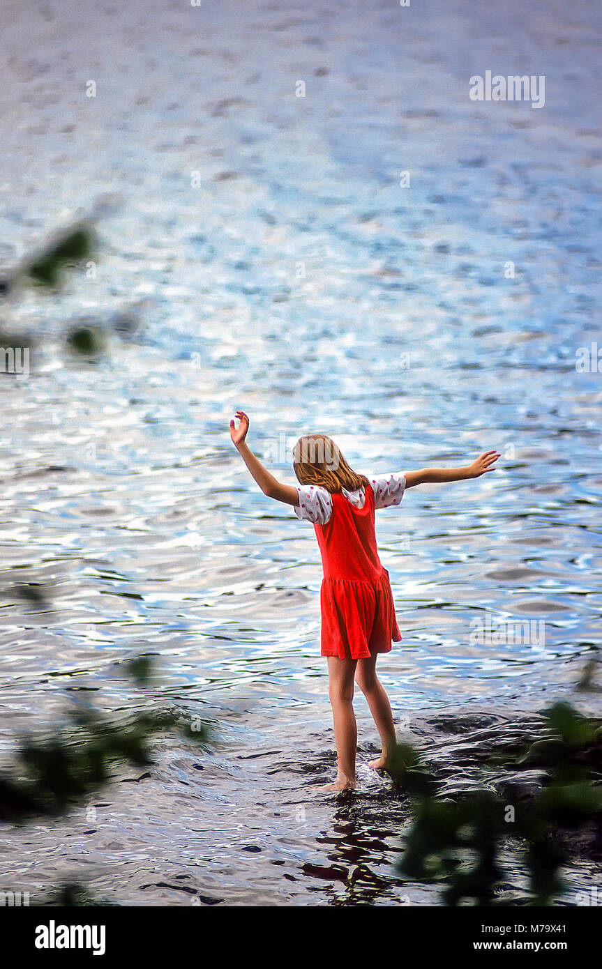 A young girl in a red dress at summer camp dips her toes into the cold water of a lake near her campground. Stock Photo