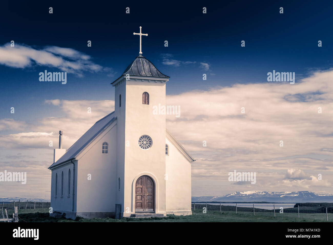 Flateyjarkirkja white lutheran church with meadow in foreground and sea  fjord with dark blue sky and mountains in the background, Flatey, Iceland Stock Photo