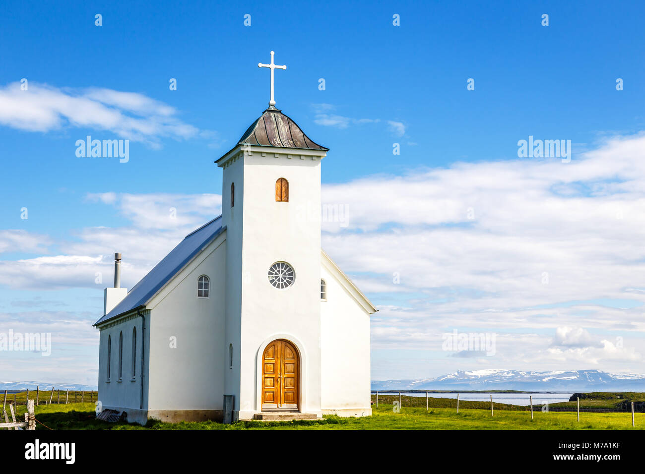 Flateyjarkirkja white lutheran church with meadow in foreground and sea  fjord with blue sky and mountains in the background, Flatey, Iceland Stock Photo
