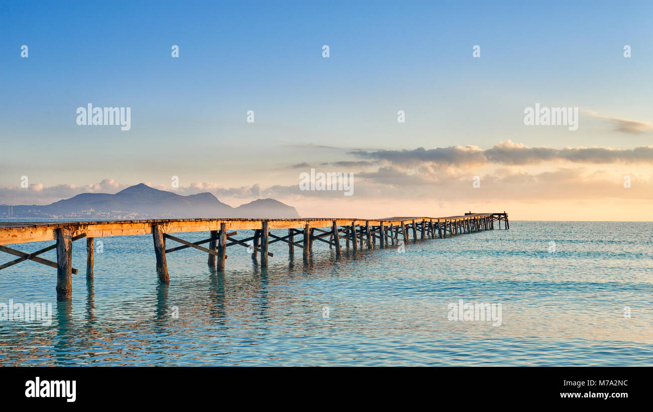 Old wooden jetty or pier stretching out into the ocean at sunset lit by the warm glow of the sun on the horizon with distant mountains over the water Stock Photo