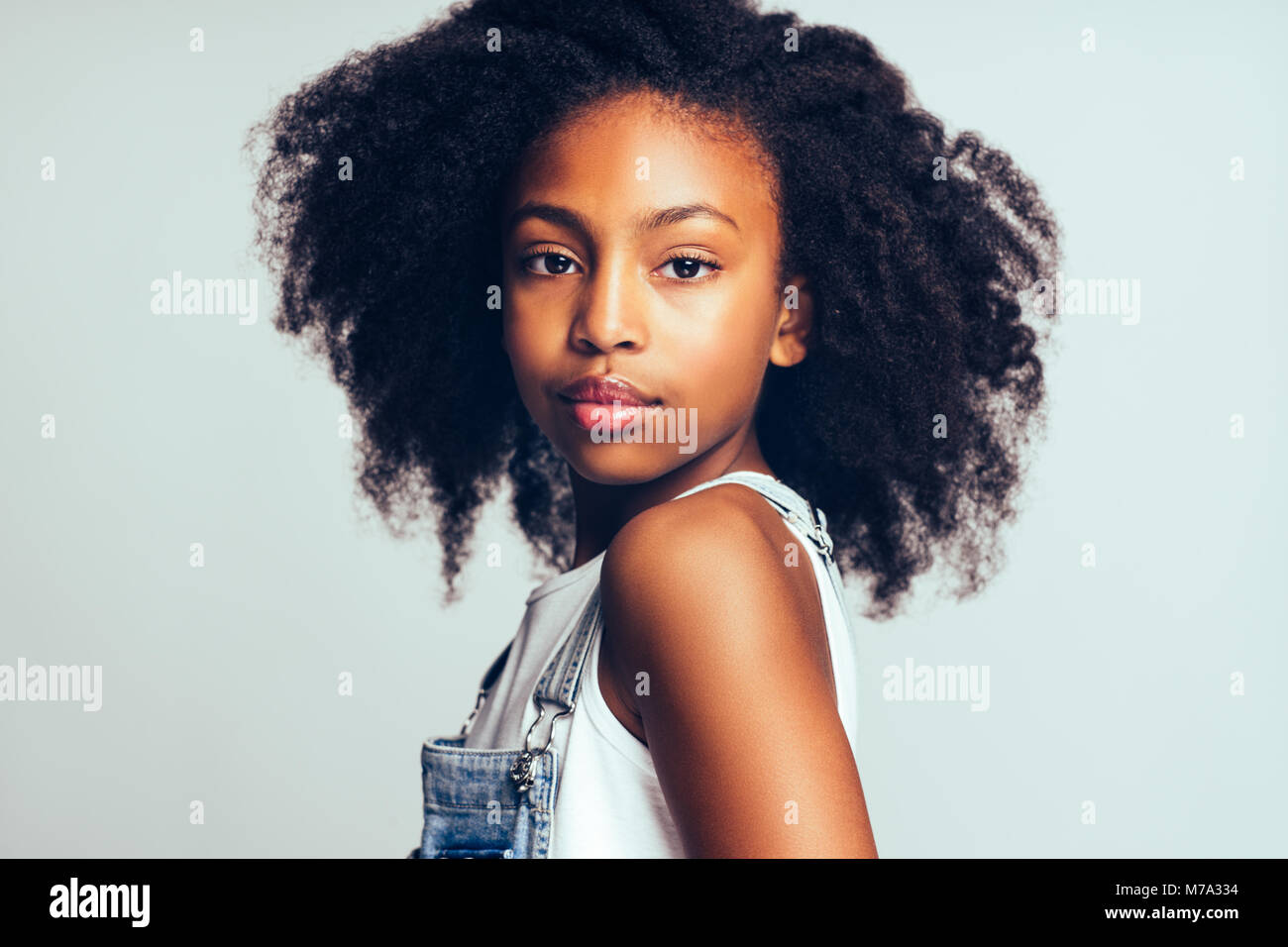 Confident young African girl with long curly hair and wearing dungarees ...