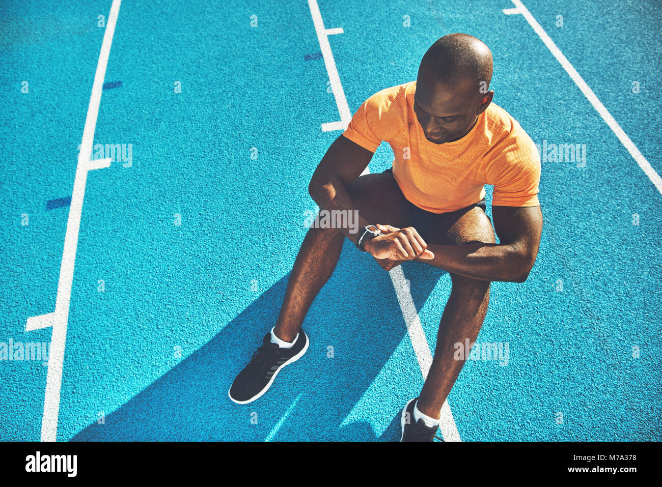 Young African male runner sitting alone on the lanes of a running track checking his lap time on his watch on a sunny day Stock Photo
