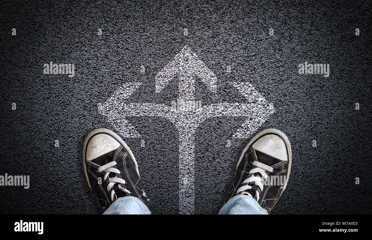 A teenager in jeans and canvas shoes standing on asphalt road with three-way arrow in the center and copy space. Concept of standing at the crossroads Stock Photo