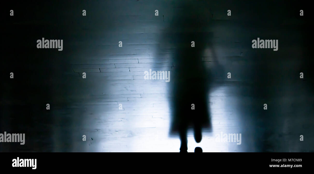 Blurry dark alley in the night with silhouette and shadow of a woman in long coat and high heels walking away Stock Photo