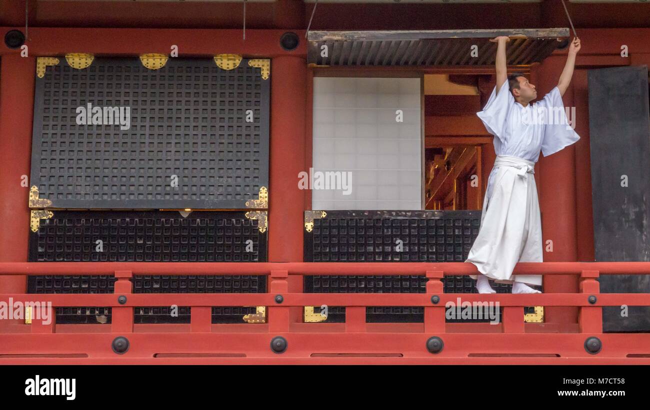 Shinto priest dressed in white robes standing on Tsurugaoka Hachimangū shrine railing closing large wooden window in preparation of the closing. Stock Photo