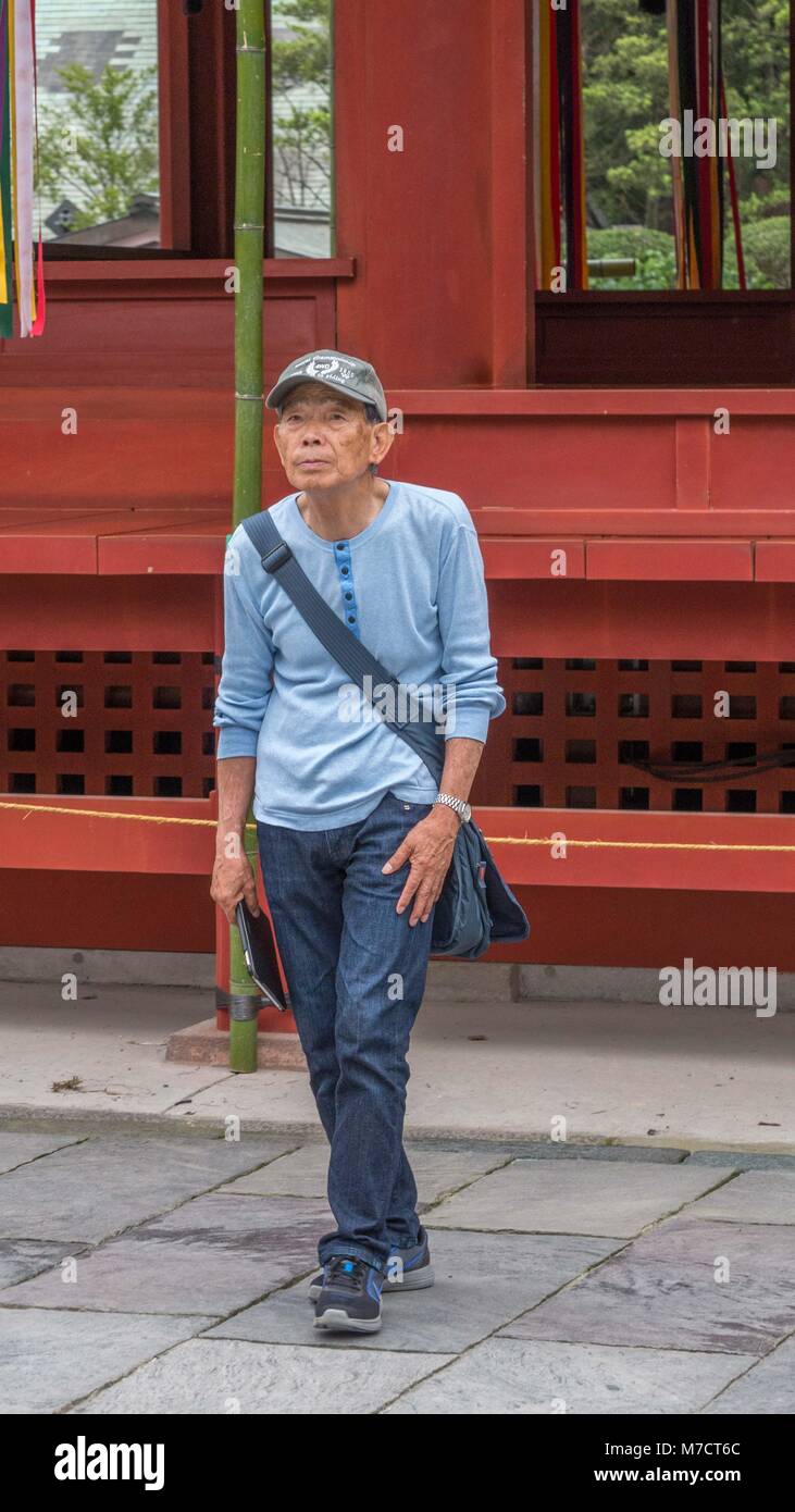Older Japanese man dressed in blue jeans and light blue shirt visiting Tsurugaoka Hachimangū shrine in Kamakura Japan red shrine in background. Stock Photo