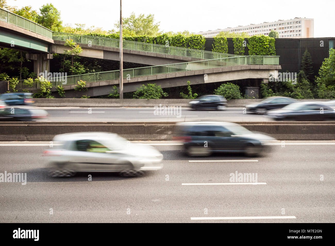 A highway with cars passing under a pedestrian footbridge, behind a noise barrier covered with vegetation, and residential building in the background. Stock Photo