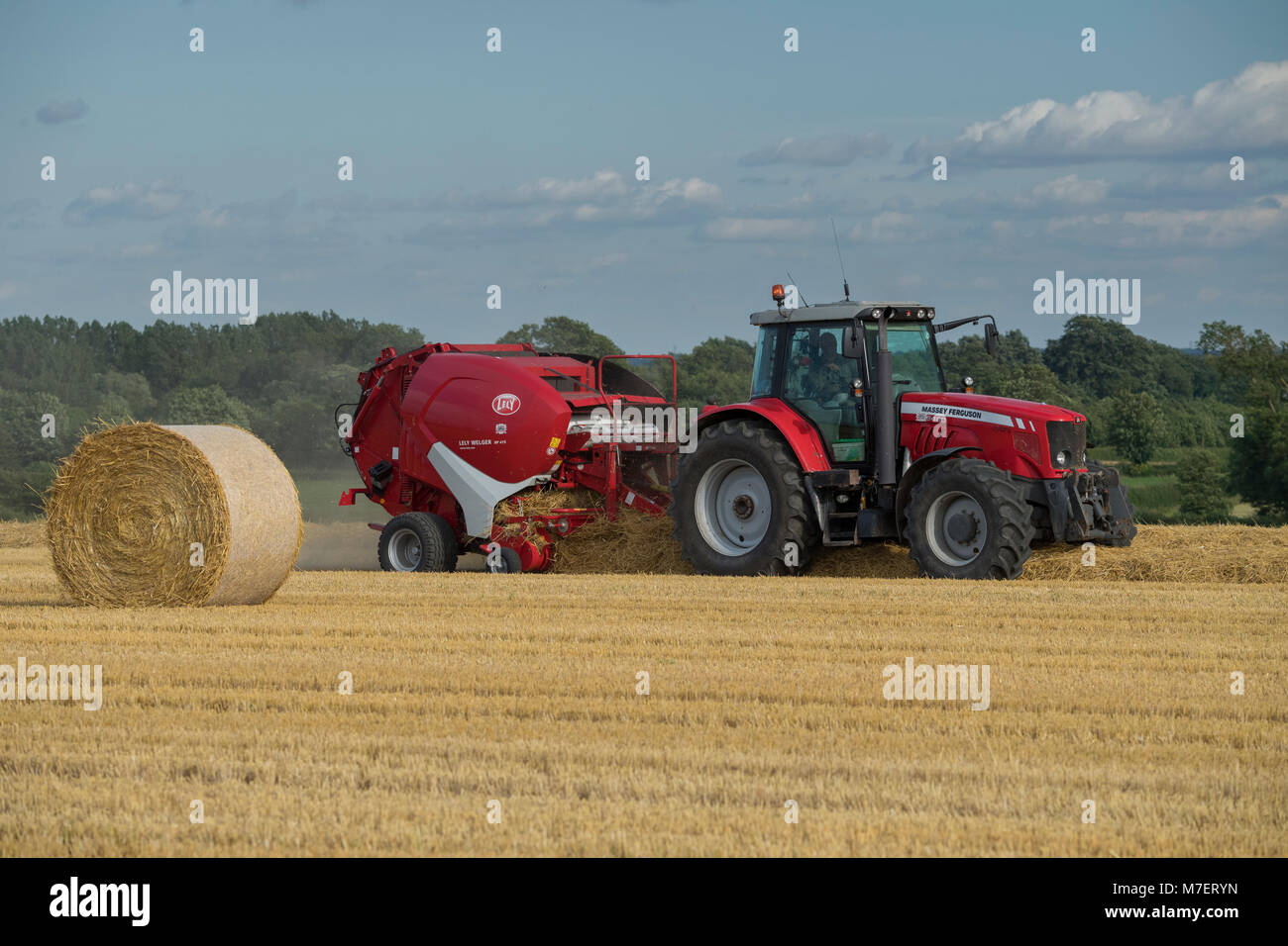 Baling straw in farm field, a farmer works & drives red tractor pulling round baler & passing large bale - Whixley, North Yorkshire, England, UK. Stock Photo