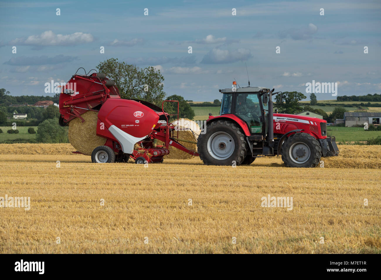 Baling straw in farm field, farmer works & drives red tractor pulling round baler (just releasing large bale) - Whixley, North Yorkshire, England, UK. Stock Photo