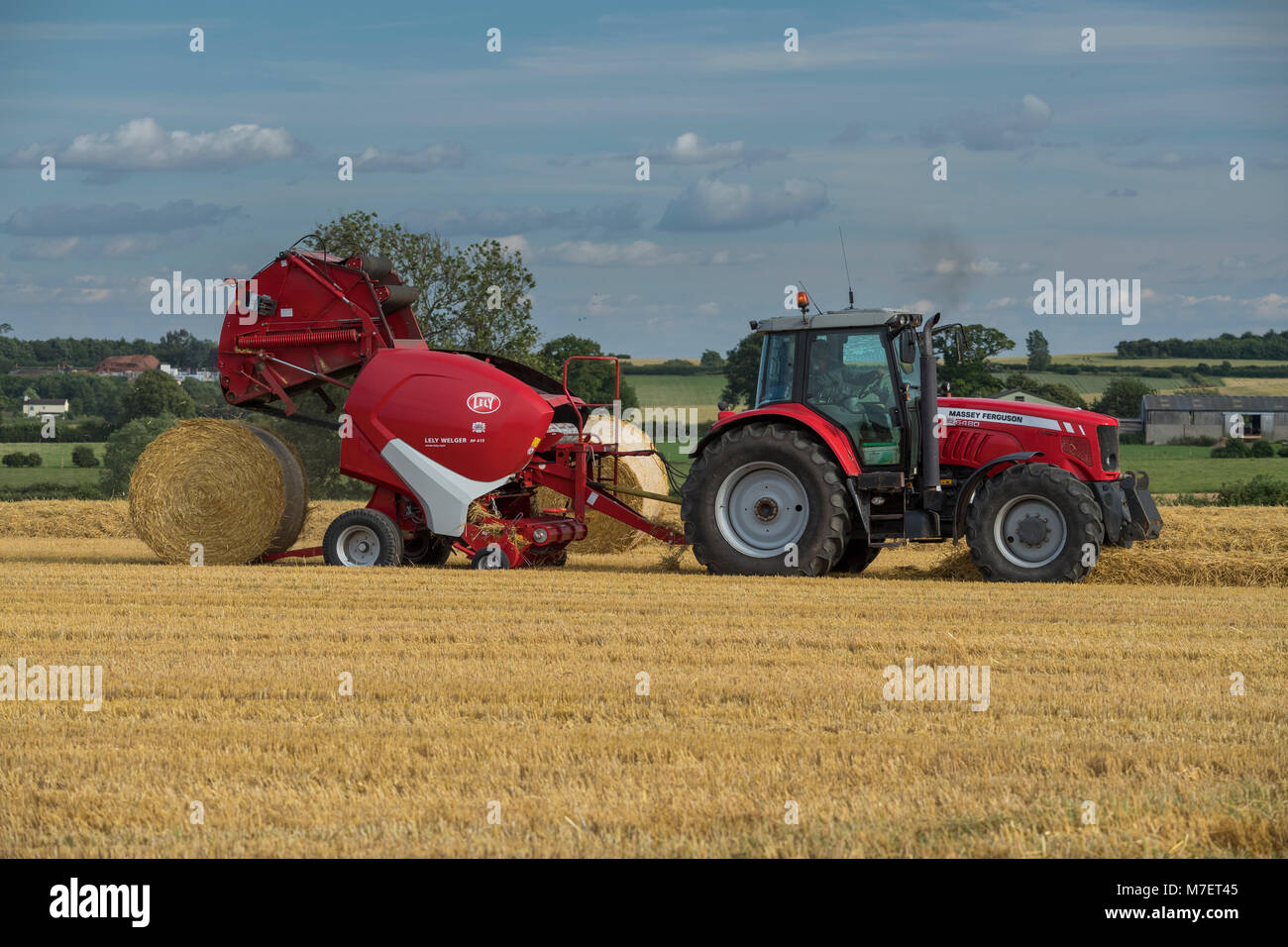 Baling straw in farm field, farmer works & drives red tractor pulling round baler (large bale just released) - Whixley, North Yorkshire, England, UK. Stock Photo
