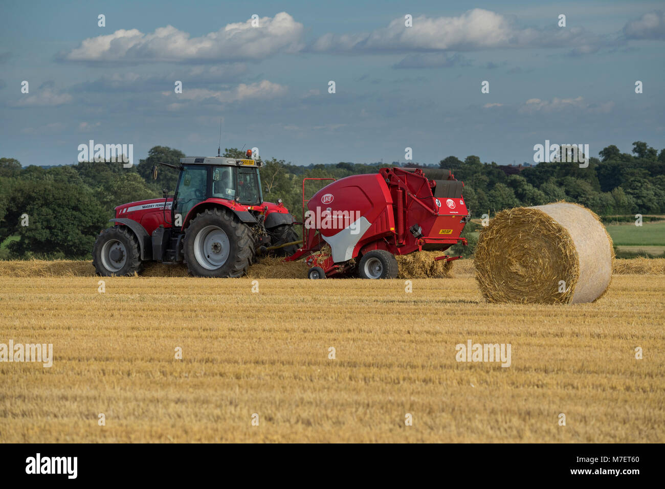 Baling straw in farm field, a farmer works & drives red tractor pulling round baler & passing large bale - Whixley, North Yorkshire, England, UK. Stock Photo
